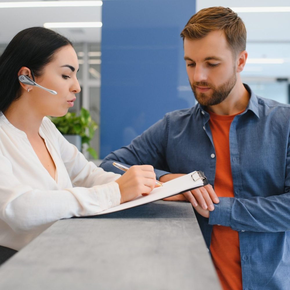The employee of the beauty salon meets the client in the reception of a modern beauty salon. A man signs a paper with the consent for maintenance. The woman smiles at him.